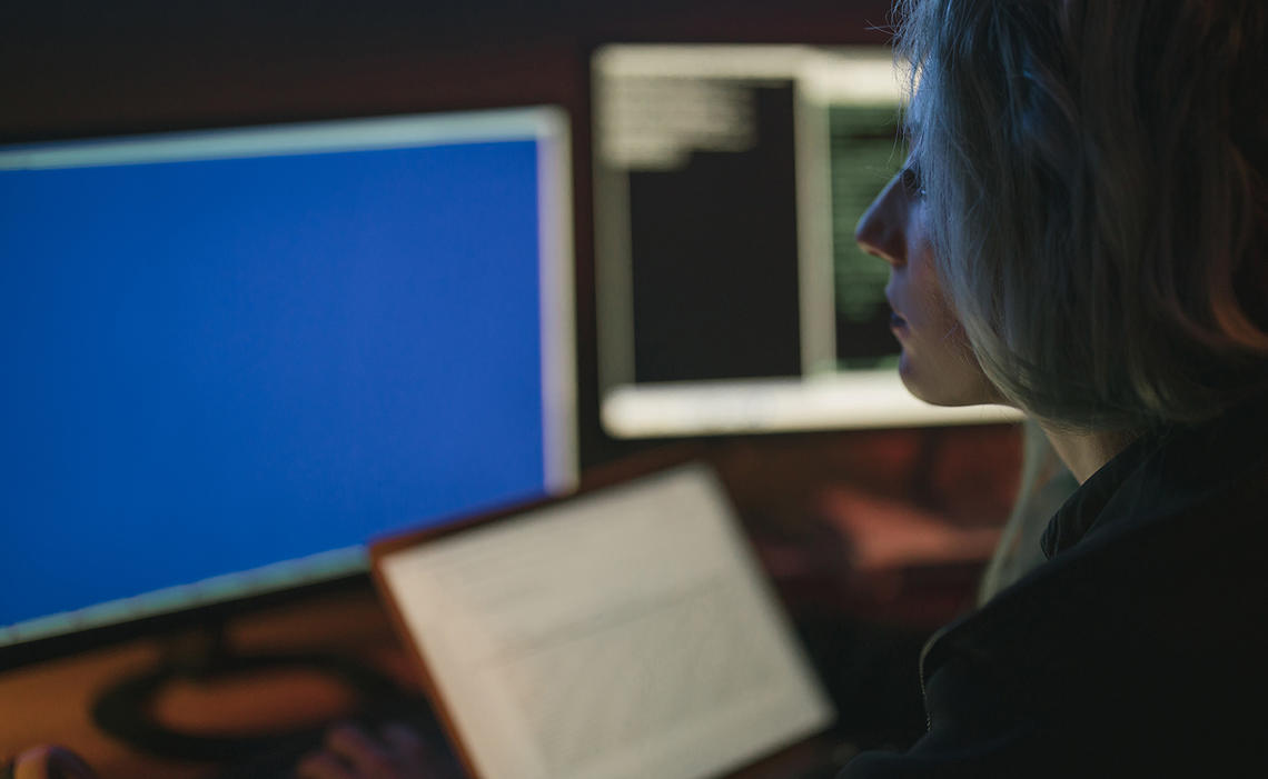 Woman sitting in a dark room with two monitors, one blue, one with coding, and a laptop