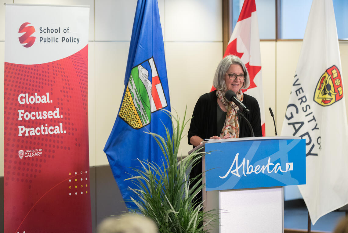 A woman stands behind a podium with Albertan, Canadian and UCalgary flags behind her