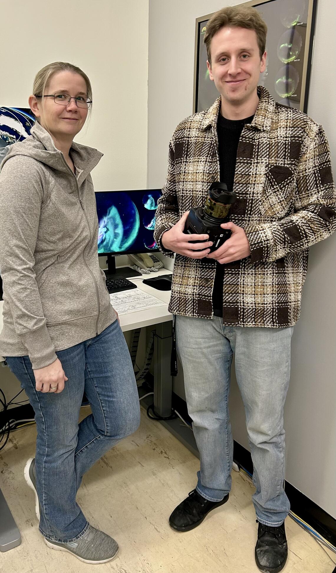 A woman and man, who is holding a camera, stand in front of a computer