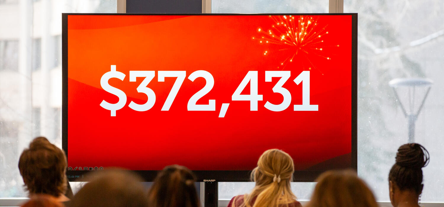 People face away from the camera looking at a large screen with a red background and the number '$372,431' on it
