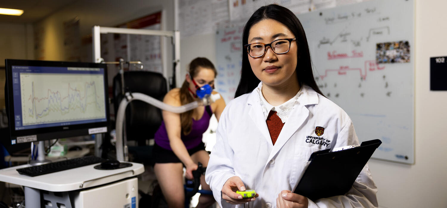 A woman wearing a lab coat and glasses smiles at the camera while another works out behind her
