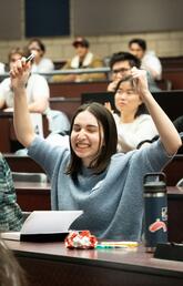 In a classroom a girl wearing a blue sweater with dark brown hair smiles with her arms in the air